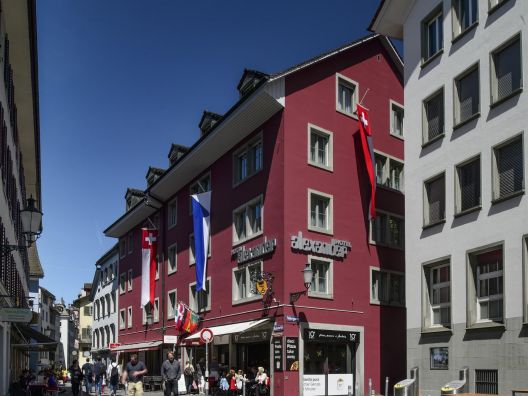 Das Hotel Alexander von aussen, ein stattliches Gebäude in roter Farbe. Aus den Fenstern hängen Fahnen der Stadt Zürich und der Schweiz.